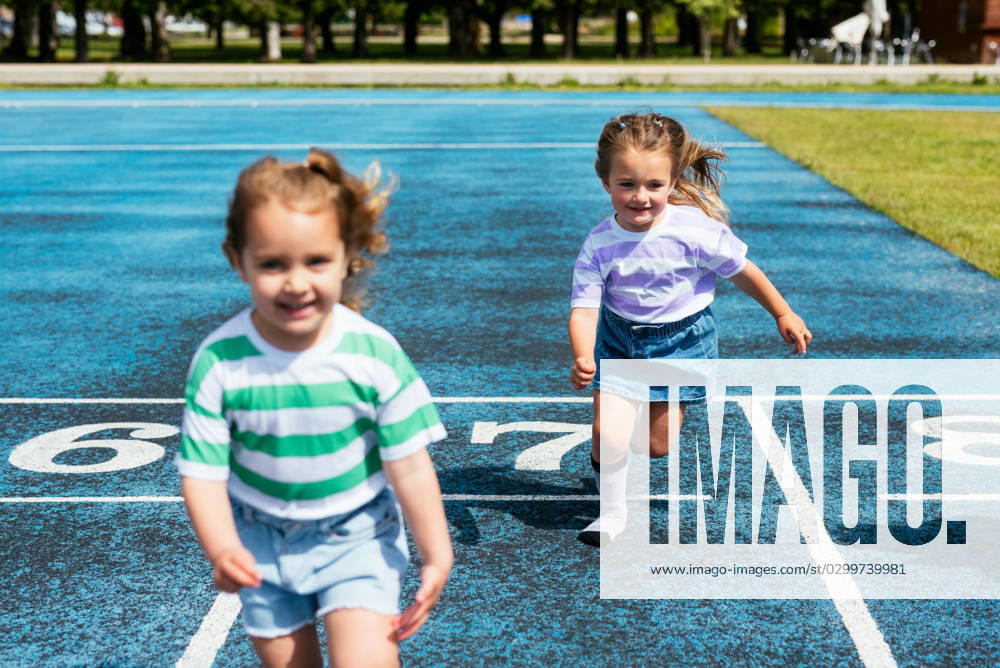 IMAGO Creative: Sommer, Sommertage im Stadtpark MR_ID14992_14133_SofiaVazquezEsmerode.jpg, MR_ID14992_14134_AdrianaMoralesPineiro.jpg Cute little girls in casual clothes smiling and running on stadium track during outdoor sports training at playground of city park, Model released AidaMartinez_ID14992_458079_011