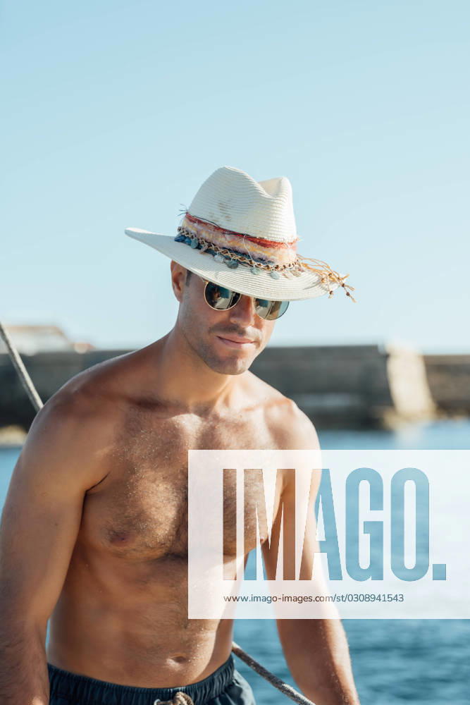 Young fit shirtless man in swimming shorts and hat standing on sailboat enjoying summer vacation against sea and cloudless blue sky Model Released AlvaroMontero_ID16707_510355_005