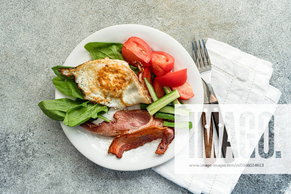 From above of balanced breakfast arrangement on a white plate with fried egg, crispy bacon, fresh spinach leaves, sliced cucumber and wedged tomatoes served with cutlery on grey textured surface Model Released AnnaBogush_ID17558_531810_002