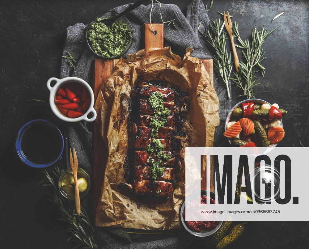 Roasted BBQ ribs with homemade pesto, pickled vegetables, sauces and rosemary on dark concrete kitchen table. Flavorful seasoned meat for dinner. Top view. Model Released Vicuschka_ID16617_505418_009
