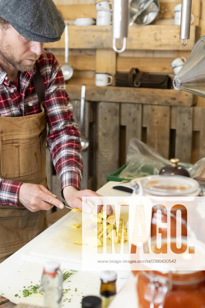 IMAGO Creative, Food, Trend, Meal Prep Crop focused chef in apron and beret standing in a rustic kitchen attentively dicing potatoes on a white cutting board with a background of hanging utensils and jars of homemade preserves Model Released JuanAizpuru_ID18273_546351_022