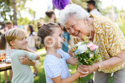 Garden birthday party for senior lady. Beautiful senior birthday woman receiving flowers from granddaughter and grandson. birthday woman senior flowers gifts garden birthday party beautiful woman lady receiving gift bouquet friends family grandmother grandchildren celebration joy happiness presents outdoors event festivity gathering special occasion surprise smiles enjoyment togetherness love affection decorations cake memories balloons laughter congratulations wishes entertainment bonding delight cheerful atmosphere friendship gratitude copy space gray hair pearls