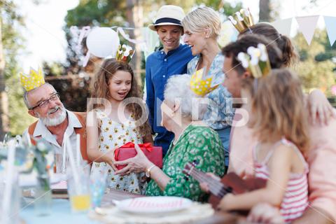 Garden birthday party for senior lady. Beautiful senior birthday woman receiving gift from granddaughter, family and friends. Birthday celebration with paper crowns. birthday woman senior birthday party garden presents gift hugging celeration gifts beautiful woman lady receiving embrace smile friends family grandmother grandson celebration joy happiness outdoors event festivity gathering special occasion surprise smiles enjoyment togetherness love affection decorations cake memories balloons laughter congratulations wishes entertainment bonding delight cheerful atmosphere friendship gratitude copy space gray hair pearls