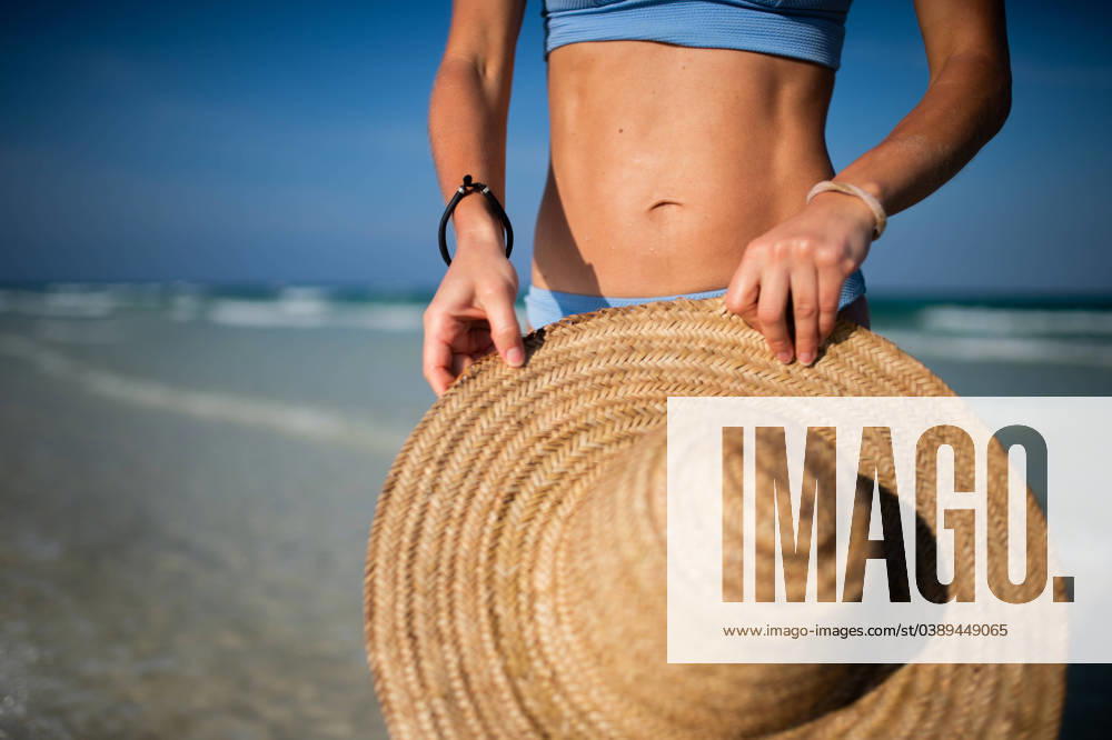 Close up of women s midsection in bikini on the sand beach near the sea. Close up of beautifuly shaped women s midsection, belly, stomach on the beach near the sea. Detail shot of woman in blue bikini on beach with straw hat in hand with copy space. Sandy beach and crystalline sea of Mnemba beach in Zanzibar. Early pregnancy on vacation. Concept of beach summer vacation. model released