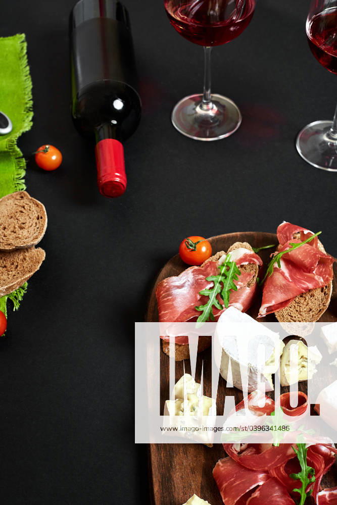 Prosciutto, salami, baguette slices, tomatoes and nutson rustic wooden board, two glasses of red wine over black background Cheese and meat appetizer selection. Prosciutto, salami, baguette slices, tomatoes and nutson rustic wooden board, two glasses of red wine over black background. Still life