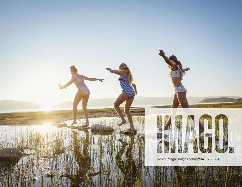 Girls (10-11, 12-13, 14-15) walking on stepping stones in lake at sunrise , 20.12.2023, model released, TTBB_ti0007_02286.jpg, Pre-Adolescent Child,Girls,10-11 Years,12-13 Years,Teenager,Teenage Girls,14-15 Years,Outdoors,Sunrise,Horizontal,Arms Raised,Walking,Sister,Family,Sun,Sunlight,Clear Sky,Summer,Landscape,Scenics,Tranquility,Lake,Lakeshore,Vacations,Rock,Vitality,Reeds,Ba