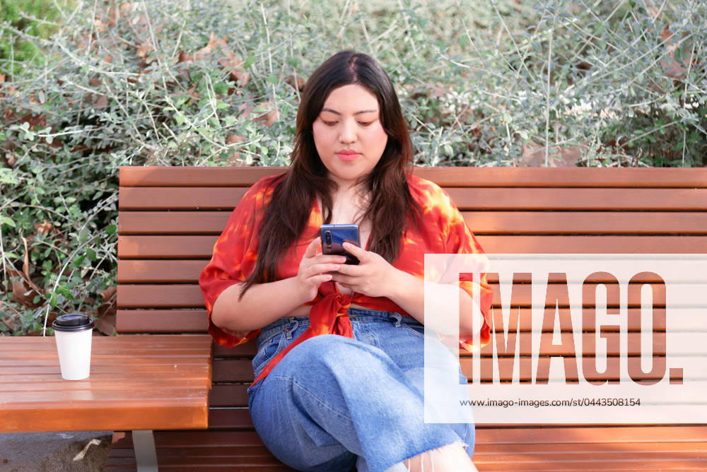 IMAGO Creative: Sommer, Sommertage im Stadtpark A young woman sits on a wooden bench in an urban park, engrossed in her phone, with a coffee cup beside her. Model Released NataliaPenado_ID20207_597968_003