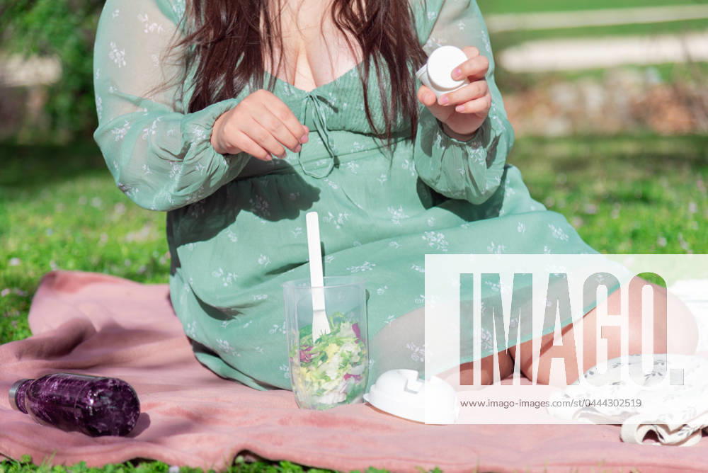 A woman in a green dress is seated on a pink blanket outside, preparing to eat a fresh salad from a clear container, with a sunny backdrop. Model Released NataliaPenado_ID20535_606443_014