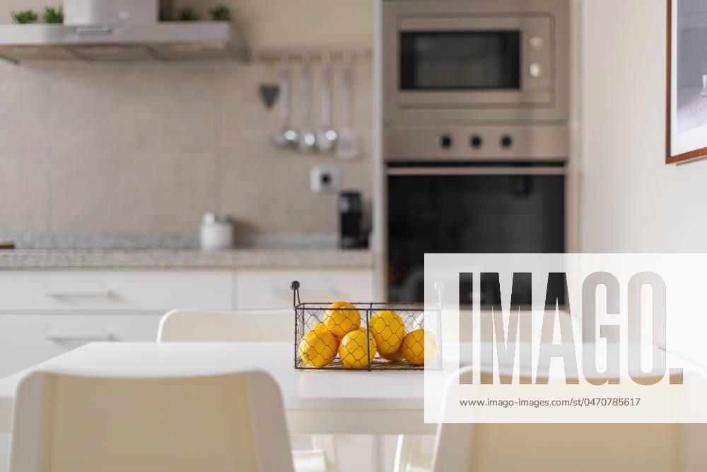 Metal basket with ripe yellow lemons on a white dining table. Blurred background of kitchen furniture and electronic equipment made of brushed durable metal along the wall. Metal basket with ripe yellow lemons on a white dining table. Blurred background of kitchen furniture and electronic equipment made of brushed durable metal along the wall