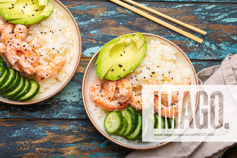 Two white ceramic bowls with rice, shrimps, avocado, vegetables and sesame seeds and chopsticks on colourful rustic wooden background top view. Healthy asian style poke bowl Two white ceramic bowls with rice, shrimps, avocado, vegetables and sesame seeds and chopsticks on colourful rustic wooden background top view. Healthy asian style poke bowl.