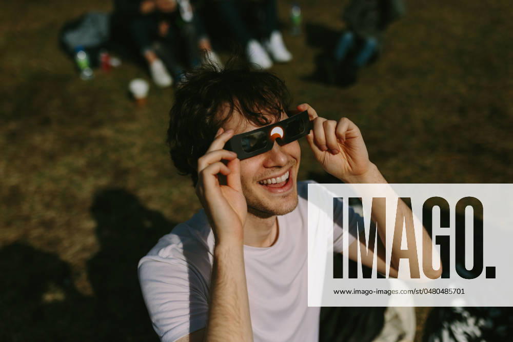 IMAGO Creative: Sommer, Sommertage im Stadtpark A cheerful young man enjoys using solar eclipse glasses in a sunlit park, surrounded by a relaxed group of friends casually gathered in the background Model Released CamilleGirard_ID21463_631383_010