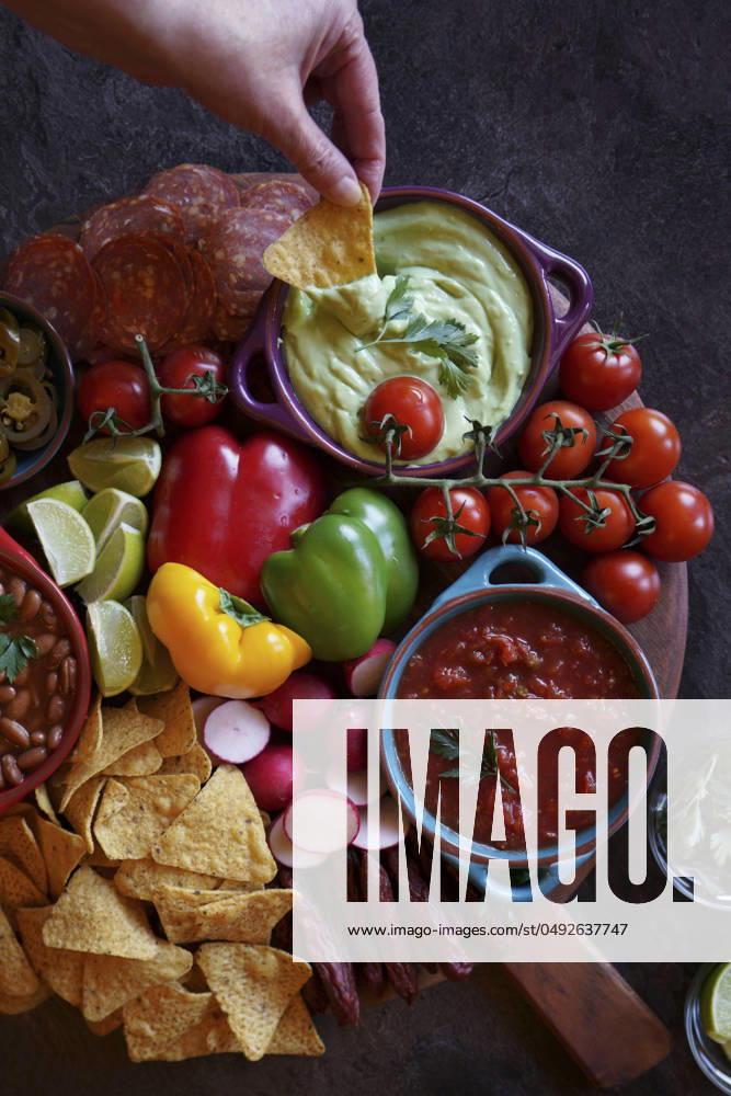 Mexican theme charcuterie board grazing platter with dips, salsa, beans, jalapenos, chorizo, and tortilla chips. Hand dipping guacamole. Mexican theme charcuterie board grazing platter with dips, salsa, beans, jalapenos, chorizo, and tortilla chips. Hand dipping guacamole.