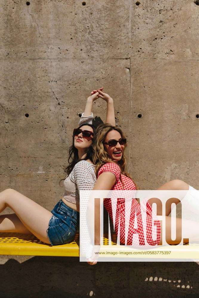 IMAGO Creative: Sommer, Sommertage im Stadtpark Two cheerful young women having fun while sitting back-to-back on a yellow bench in a park, both dressed in summer outfits and sunglasses, capturing the essence of friendship and joy Model Released CamilleGirard_ID21698_636447_015