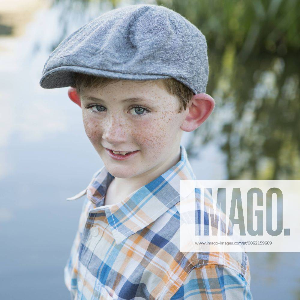 A young boy wearing a checked shirt and cloth cap with a large brim standing on a riverbank.