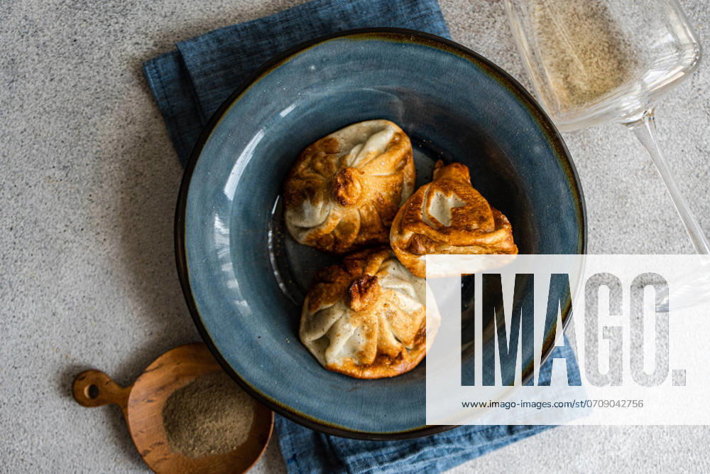 IMAGO Creative, Food, Dumplings A close-up view of traditional Georgian khinkali dumplings on a textured blue ceramic plate, accompanied by a wooden scoop of spices and a clear wine glass, set against a gray tabletop Model Released AnnaBogush_ID22388_654123_002