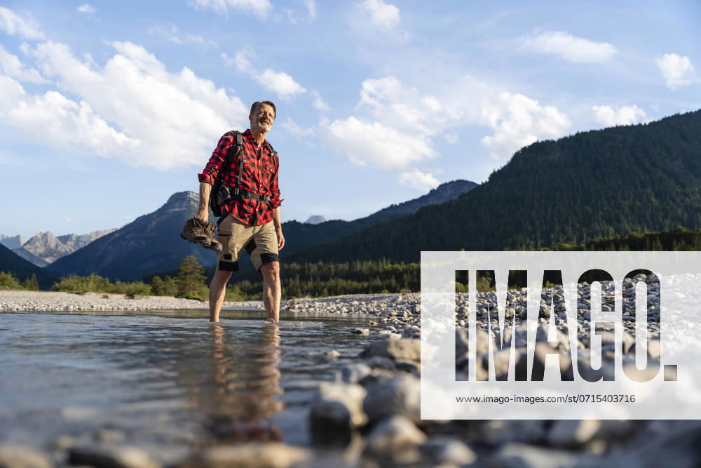 Senior man hiking through a rocky stream in the mountains with a backpack, flannel shirt, and shorts on a sunny day. model released, Symbolfoto, DIGF21510