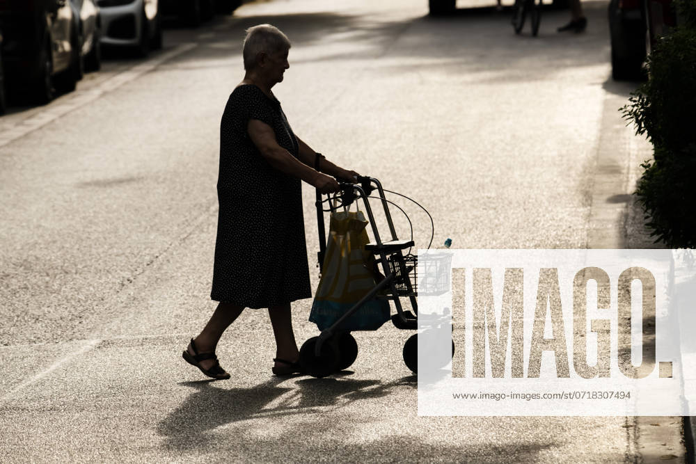 Old woman with rollator walking in the city in the summer heat, July 2024 Germany, Munich, July 2024, old woman with rollator walking in the heat, in the summer heat there is a risk of dehydration, especially for older people, senior citizen has taken a water bottle with her, strong sunlight, back