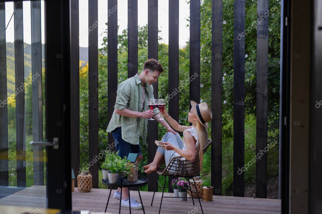 A young couple clinking with wine outdoors, weekend away in tiny house in countryside, sustainable living.