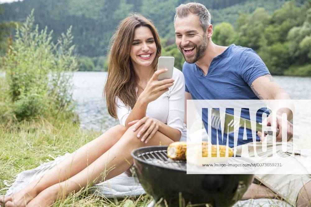 Happy couple with cell phone having a barbecue at lakeside model released Symbolfoto FMKF002847