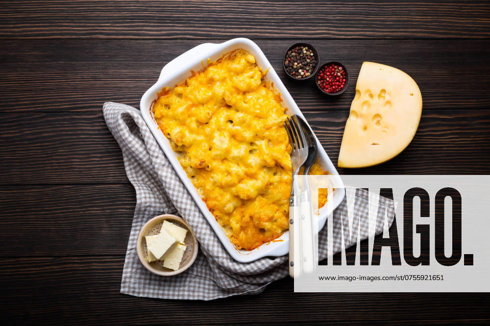 Nahaufnahme von gebackenen Maccaroni und Käse in weißer Kasserolle auf rustikalem Holzhintergrund, mit Gewürzen, Butter, Löffel und Gabel, Ansicht von oben. Nudeln mit Käsesauce, amerikanische englische Hausmannskost Close-up of baked mac and cheese in white casserole on rustic wooden background, with seasonings, butter, spoon and fork, top view. Pasta with cheesy sauce, American English comfort food