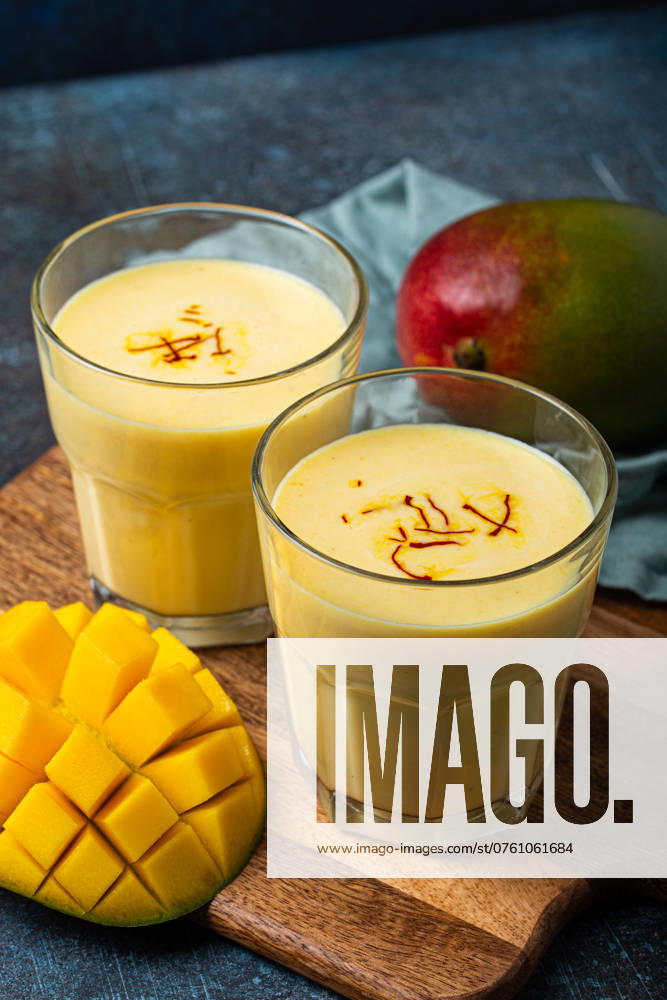 Gesundes indisches Ayurveda-Getränk Mango Lassi in zwei Gläsern auf rustikalem Betontisch mit frischer, reif geschnittener Mango, gelbes Mischgetränk aus Mangofrucht, Joghurt oder Milchquark und Gewürzen Healthy Indian Ayurveda drink mango lassi in two glasses on rustic concrete table with fresh ripe cut mango, yellow blended beverage made of mango fruit, yoghurt or milk curd and spices