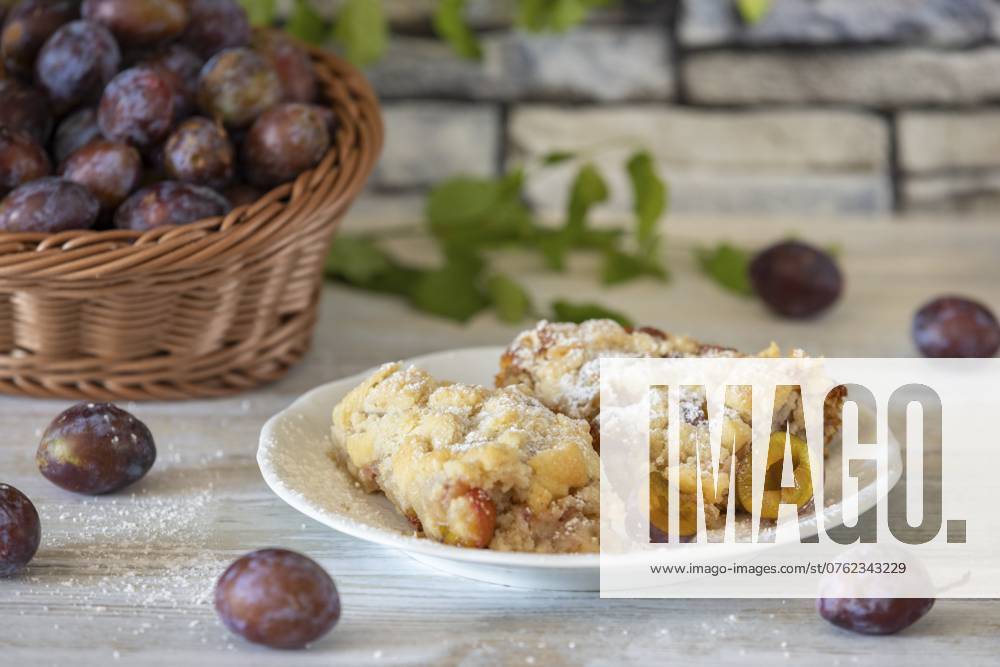 IMAGO Creative, Herbst, Food, Pflaumenkuchen Zwetschgenkuchen auf einem Teller neben Korb mit frischen Zwetschgen im Hintergrund, auf einem weißen rustikalen Tisch Plum cake on a plate next to a basket with fresh plums in the background, on a white rustic table
