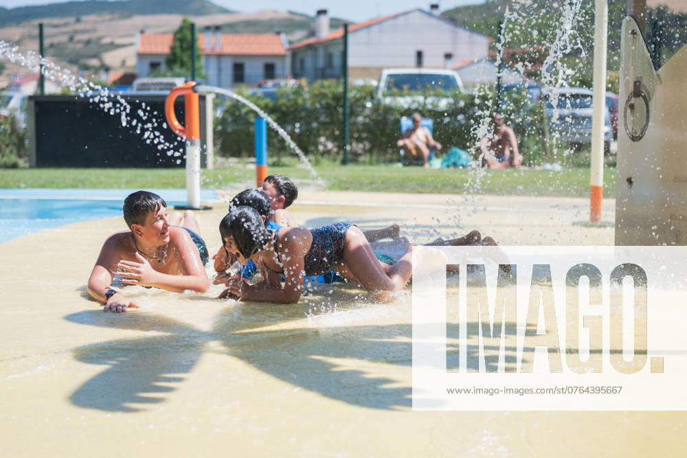 IMAGO Creative, Jahreszeit Sommer, Hitze Kids playing under a water spray in a pool area on a hot summer day, cooling off and splashing. The scene captures children lying prone and discussing excitedly, some looking at each other, engaged in lively conversations. Model Released HodeiUnzueta_ID23816_691396_004