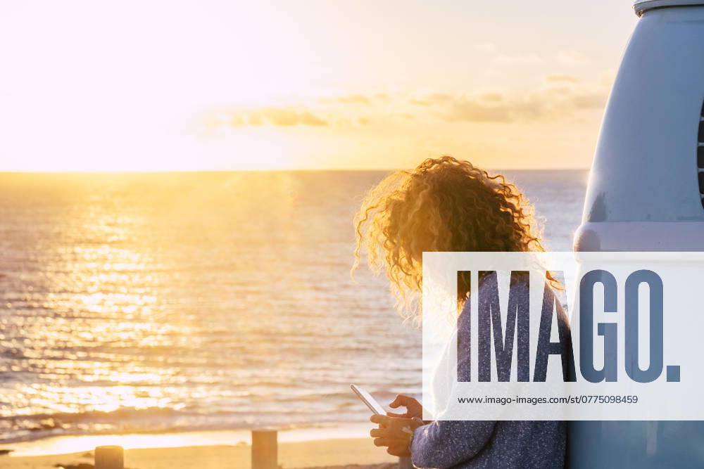Woman using smart phone by beach at sunset in Fuerteventura, Canary Islands , 04.07.2019, model released, TTBB_ti0564_00080.jpg, Color Image,Horizontal,Focus On Foreground,Selective Focus,Outdoors,Sunset,Dusk,Fuerteventura,Canary Islands,Spain,Travel Destinations,Travel,One Person,People,40-44 Years,Mid Adult,One Mid Adult Woman Only,Only Women,Adult,Mid Adult Women,Caucasian Appearance,Personal Land Vehicle,Camper Van,Road Trip,Transportation,Getting Away From It All,Tourist,Beach,Sand,Coastline,Sea,Water,Blond Hair,Curly Hair,Sweater,Holding,Smart Phone,Communication,Sunbeam,Sunlight,Text Messaging,Mobile Phone,Wireless Technology,Cloud,Leaning