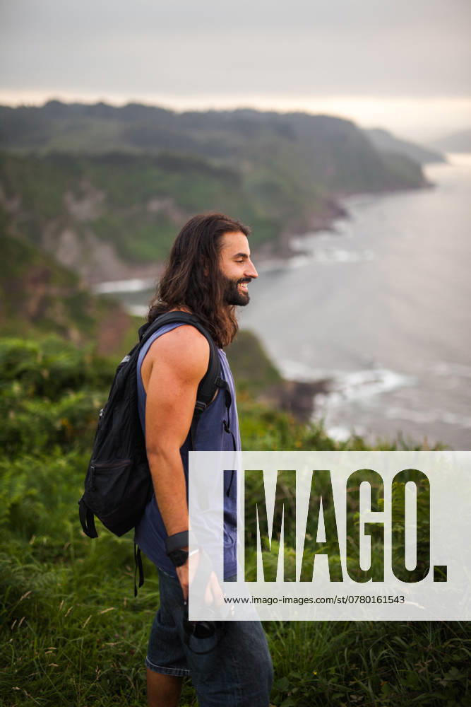 A joyful male traveler with a backpack and camera experiences the lush, green landscapes and serene coastal lines of Asturias, Spain, showcasing the freedom of outdoor adventures Model Released PabloMenendez_ID23597_685263_007