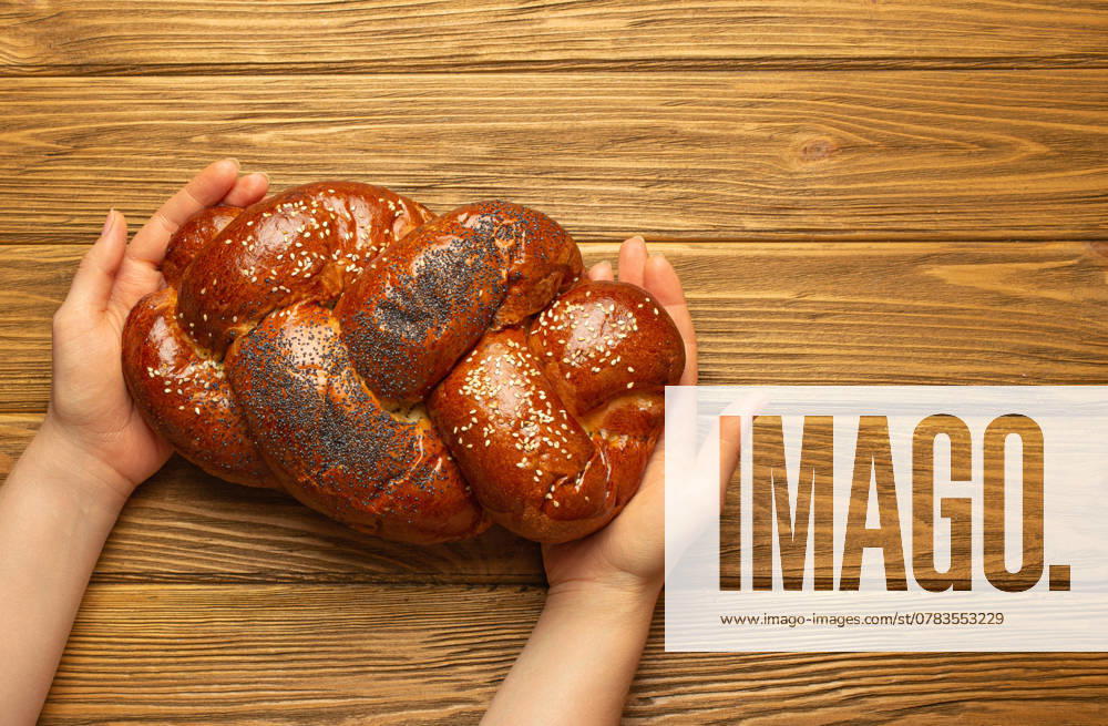Weibliche Hände halten frisch gebackenes Challah-Brot mit Mohn- und Sesamkörnern, Draufsicht auf rustikalen Holzhintergrund, traditionelle festliche jüdische Küch Female hands holding freshly baked Challah bread covered with poppy and sesame seeds, top view on rustic wooden background, traditional festive Jewish cuisine.