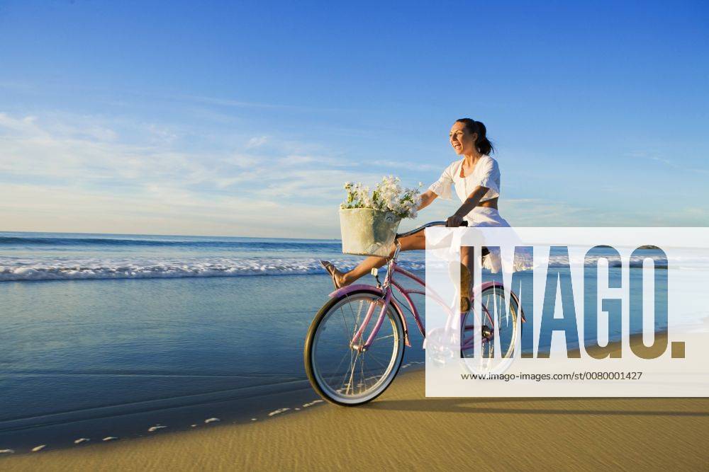 Young woman wearing a white dress riding her bike on the beach at sunset A5