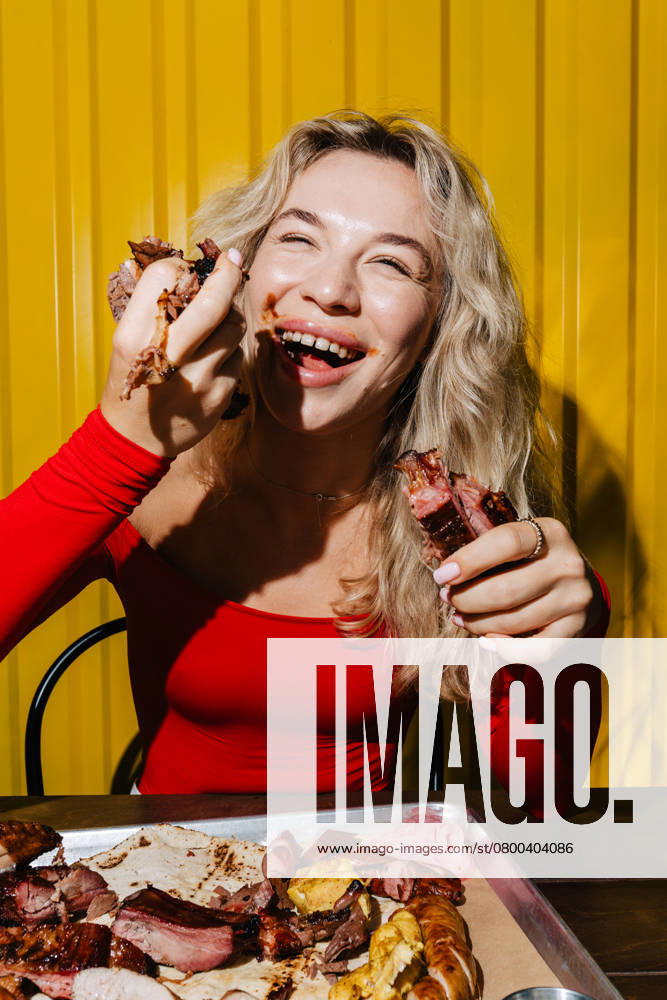 MR_ID26394_19595_MarynaMezentseva.jpg, MR_ID26394_19593_YevgeniiAkymenko.jpg, MR_ID26394_19594_KaterynaFrosiniak.jpg A happy woman in a red top enjoys eating barbecue meat with her hands. Sitting in front of a tray of grilled food, she smiles brightly, savoring the moment in a vibrant setting. Model Released Andrerako_ID26394_761141_009