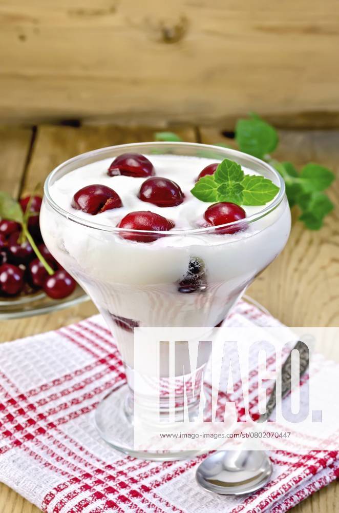IMAGO Creative, Jahreszeit Sommer, Kirschen Dicker Joghurt im Glas mit Kirsche und Löffel auf einer Serviette, Kirschen in der Untertasse, Minze auf einem Holzbrett im Hintergrund Thick yogurt in glass with cherry and spoon on a napkin, cherries in the saucer, mint on a wooden boards background