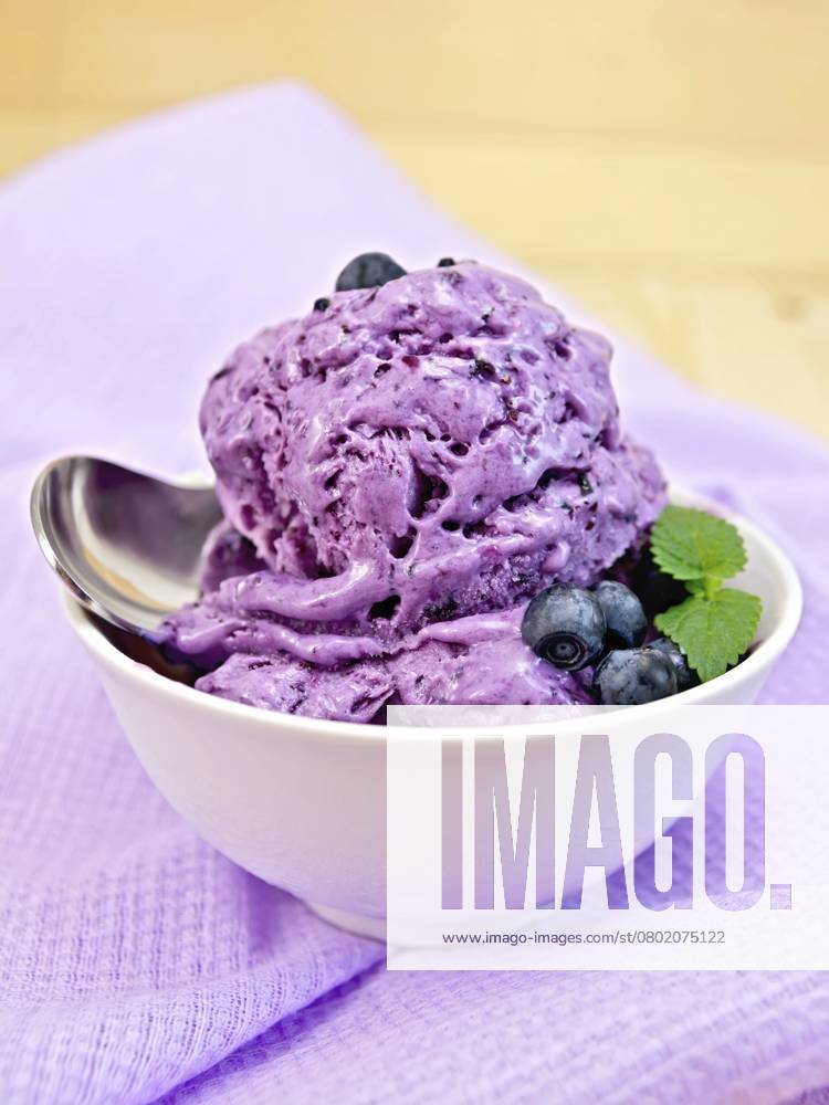 Heidelbeereis mit Minzblättern in einer Schale mit Beeren, Minze und Löffel auf einem Hintergrund aus lila Stoff und Holzbrettern Blueberry ice cream with mint leaves in a bowl with berries, mint and spoon on a background of purple cloth and wooden boards
