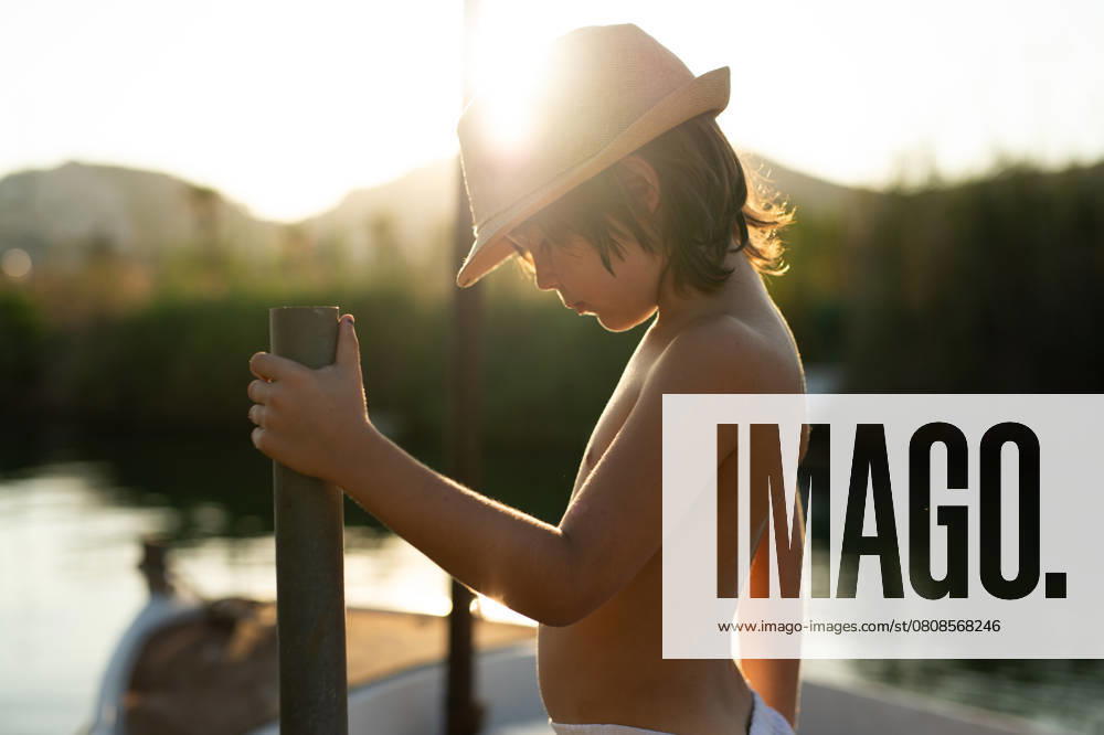 Side view of a young boy wearing a straw hat is captured leaning on a boat railing, looking down at the waters of Andratx, Mallorca. The warm sunset creates a serene backdrop emphasizing a peaceful maritime scene. Model Released MagdalenaCarli_ID27192_782031_001