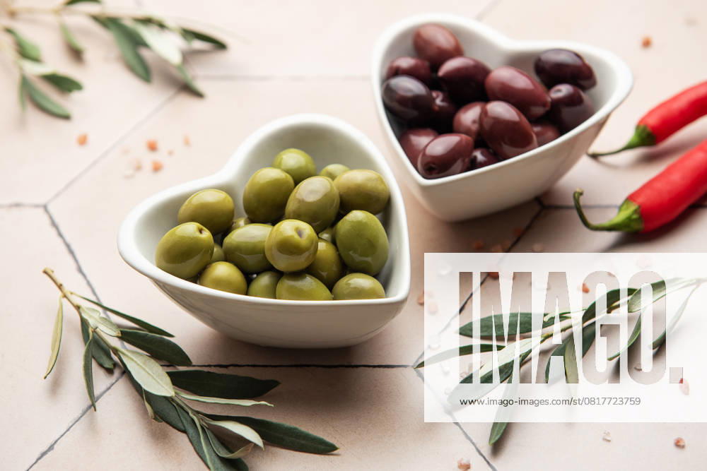 Herzförmige Schalen mit grünen und schwarzen Oliven, Olivenzweigen, Chilischoten und Himalayasalz auf gefliester Oberfläche Green and black olives served in heart shaped bowls, decorated with olive tree branches, chili peppers and himalayan salt, creating an elegant and appetizing presentation (License=RF)
