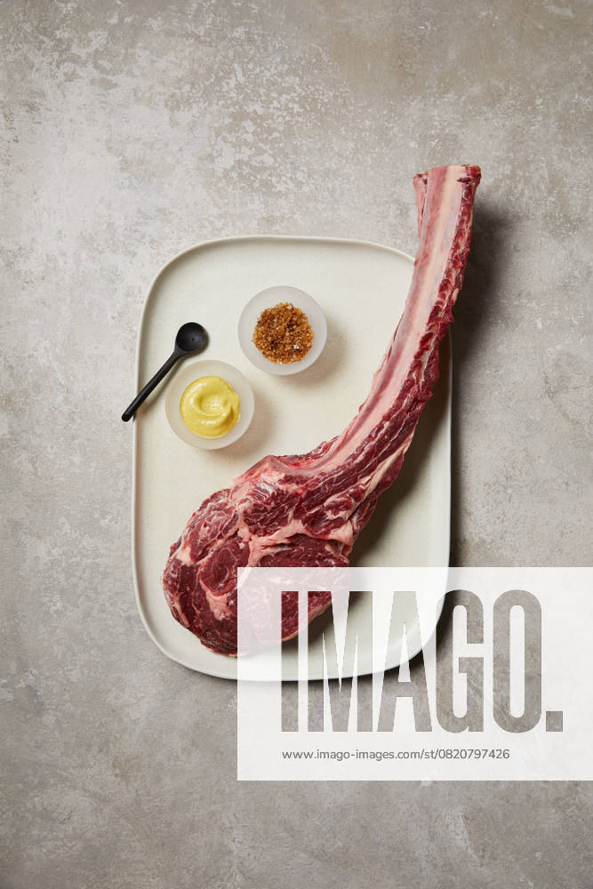 Top-down view of a raw tomahawk steak presented on a ceramic tray, accompanied by a small bowl of mustard, spices, and a black spoon. A premium cut ready for gourmet cooking Model Released GeorgiaGold_ID28577_843647_038