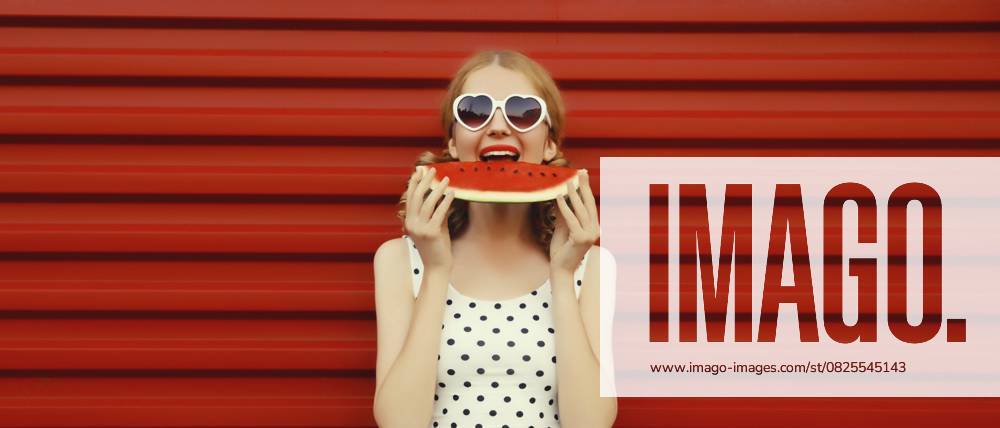 Sommerporträt einer glücklichen, lächelnden jungen Frau, die frische, saftige Früchte ißt, ein Stück Wassermelone mit einer herzförmigen Sonnenbrille auf rotem Hintergrund Summer portrait of happy smiling young woman eating fresh juicy fruits, slice of watermelon wearing heart shaped sunglasses on red background