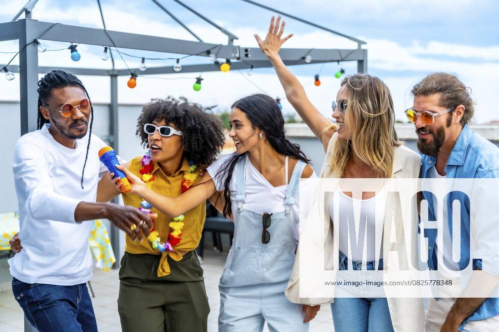 Eine Gruppe verschiedener Freunde feiert auf einem Dach, singt Karaoke, tanzt fröhlich und genießt gemeinsam eine ausgelassene Sommerparty Group of diverse friends celebrating on a rooftop, singing karaoke, dancing joyfully, and enjoying a vibrant summer party together