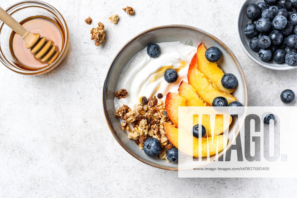 IMAGO Creative, Food, High Protein Frühstück Müslischale mit griechischem Joghurt, Pfirsich und Heidelbeeren Granola bowl for healthy breakfast on white concrete background, top view. Homemade granola with greek yogurt, peaches and blueberries. Healthy eating, seasonal summer breakfast template. (License=RF)