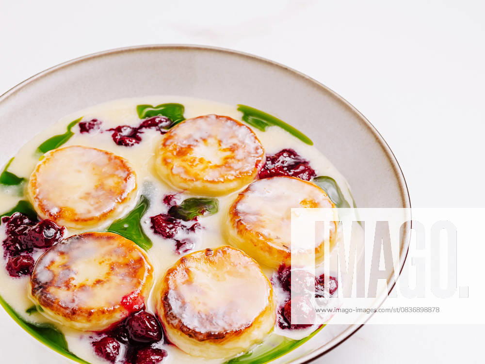 IMAGO Creative, Food, High Protein Frühstück Hüttenkäsepfannkuchen mit Sahne und Beerensauce This unique dessert features round creamy cakes topped with a glossy glaze surrounded by vibrant green herbs and red berries in a shallow bowl, presenting an elegant and delightful presentation. (License=RF)