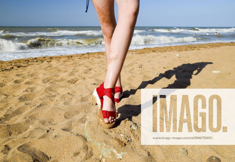 Rote Sandalen an den Beinen eines Mädchens, das auf dem Strandsand steht Red Sandals on the Legs of a Girl Standing on the Beach Sand