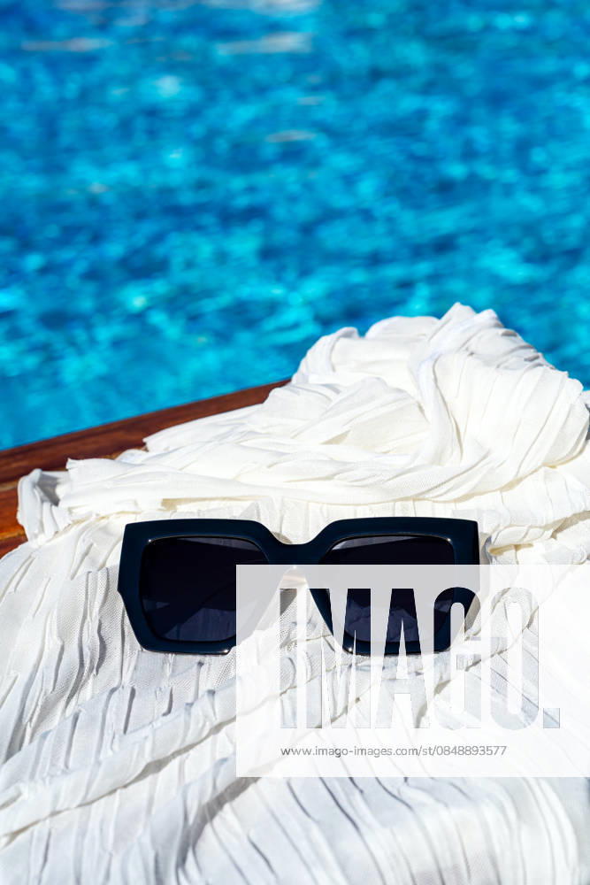 Stylish sunglasses on fabric with refreshing pool backdrop A pair of sleek black sunglasses rests on white fabric, set against a glistening blue pool backdrop. This stylish setup captures the essence of sunny relaxation and leisure. Model Released IrinaChetverikova_ID33496_1166281_001