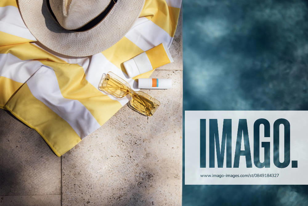 Sommer-Strandaccessoires, darunter ein Strohhut mit breiter Krempe, ein gelb gestreiftes Handtuch, eine Sonnenbrille und Sonnencreme, die auf einer Steinfläche in der Nähe eines Swimmingpools liegen. Summer beach accessories such as a straw hat, yellow striped towel, sunglasses, and sunscreen arranged on a stone surface beside a swimming pool (License=RF)