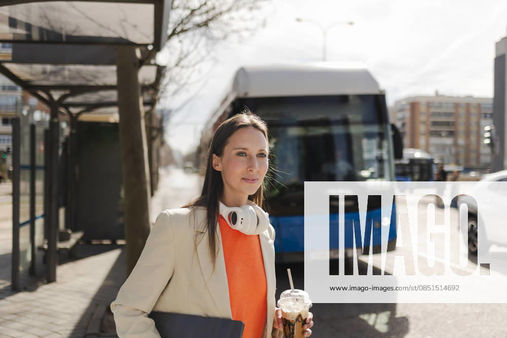 IMAGO Creative, Food, Trend, Iced Coffee Businesswoman with headphones and iced coffee waiting at urban bus stop model released, Symbolfoto, JCCMF14796