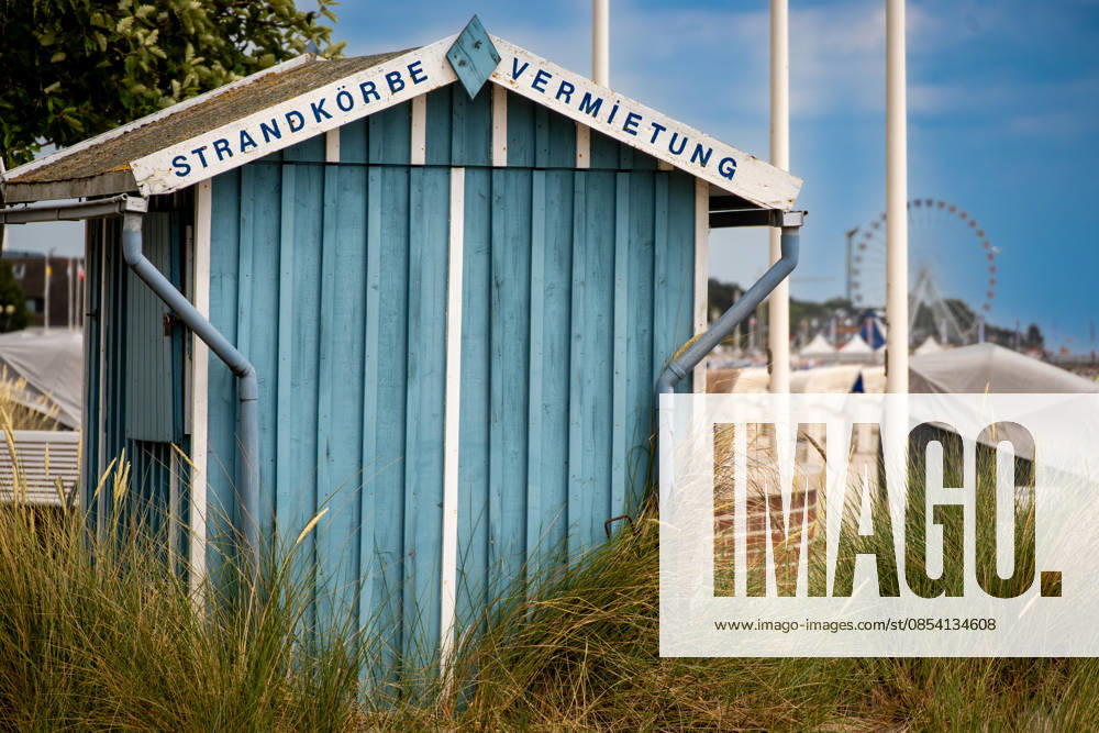 Blaue Strandkorb-Verleihhütte hinter dem Dünengras Groemitz - Schleswig-Holstein - Germany - 06-28-2025: Blue wooden hut with a beach chair rental sign stands behind dune grass, with a sandy area and a distant ferris wheel under clear sky. (License=RF)