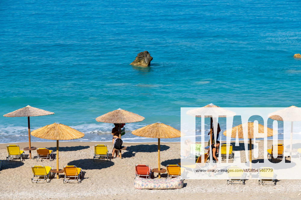Sonnenschirme und Liegestühle mit Menschen am Meer Nydri - Ionian Islands - Greece - 08-09-2025: Beach scene with straw umbrellas, yellow and orange sunbeds, people relaxing on the sandy shore, and turquoise sea with visible rock. (License=RM)