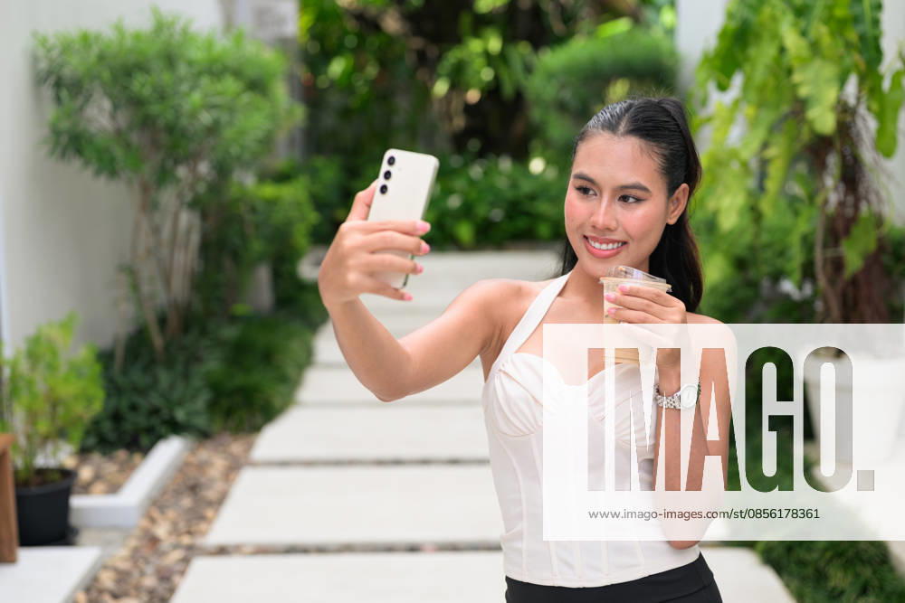 IMAGO Creative, Food, Trend, Iced Coffee Ein fröhlicher Influencer aus dem thailändischen Isan, der während der Sommerferien im Garten mit seinem Handy einen Vlog über Eiskaffee dreht Thai woman from the Isan region vlogging on a white stone walkway. The beautiful Isaan lady wears a cream corset halter blouse while holding a smartphone and iced coffee. That attractive influencer radiates pure happiness while standing outdoors in a luxury resort. Botanical backdrop showcases lush tropical foliage and dense shrubs under bright natural daylight. A stylish brunette fashion model records content for her digital audience. Ivory bodice displays ribbed textures and a structured sweetheart neckline. A fashionable subject stands amidst vibrant greenery in a serene garden environment. This g