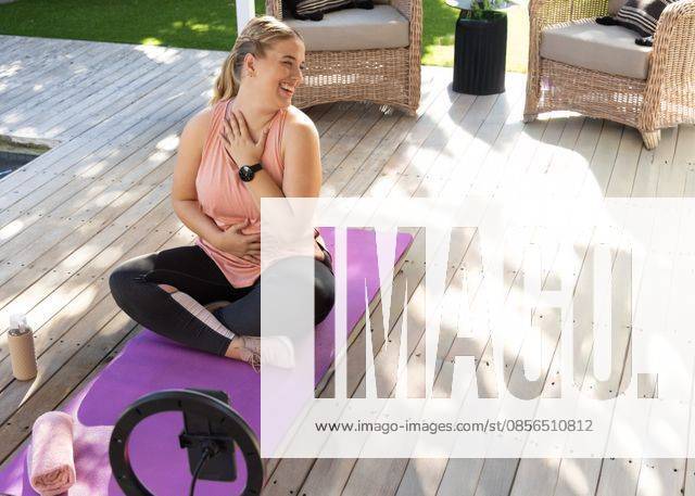 Woman sitting cross-legged on purple yoga mat on deck patio, smiling, holding chest, ring light, model released, property released, 17.04.2025 06:00:00, 13158324