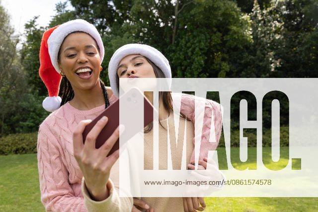 Diverse female friends posing for selfie in park holding smartphone wearing Santa hats and sweaters, model released, property released, 14.08.2025 06:00:00, 7811309